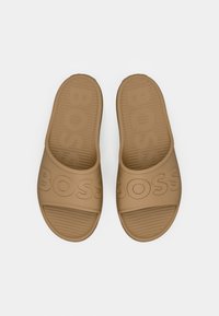 Sandales slides beige en caoutchouc, avec une semelle intérieure texturée et une inscription "BOSS" en relief sur la bride. Semelle plate sans talon.
