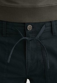 Nahaufnahme von dunkelblauen Jeans mit Metallknopf und gebundenem Kordelzug an der Taille, kombiniert mit einem gerippten dunkelgrünen Pullover.