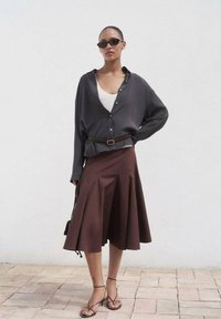 Camicia grigia button-up morbida, canotta bianca sotto, gonna plissettata marrone e sandali marroni con cinturini, abbinati a occhiali da sole scuri.
