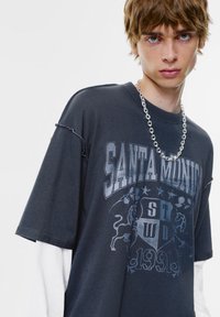 Sötétszürke, túlságosan nagy t-shirt grafikus nyomtatással, amelyen a "SANTA MONICA" felirat és egyéb részletek találhatók. Fehér hosszú ujjas póló alatt viselve.