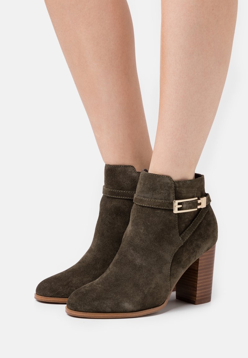 Bottes chevilles en daim marron avec talons blocs empilés en bois et détail de boucle dorée sur les jambes du modèle sur fond blanc.
