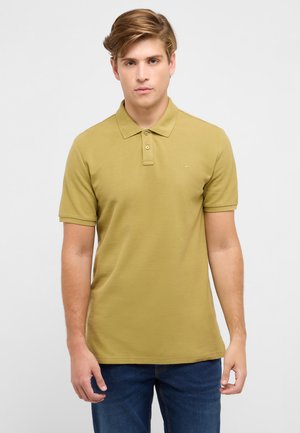 Młody mężczyzna o jasnobrązowych włosach, ubrany w musztardowożółtą koszulkę polo z krótkim rękawem i ciemnoniebieskie dżinsy, na jasnym, jednolitym tle.