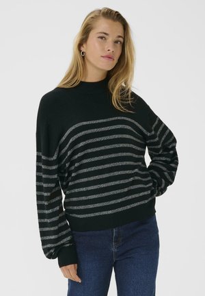 Jonge vrouw met lang blond haar, gekleed in een zwart sweater met zilveren strepen en donkerblauwe jeans, staande met één hand in haar zak tegen een effen achtergrond.