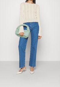 Cremfarbener Strickpullover mit offenen Mustern, blaue hochtaillierte Jeans, mintgrüne Handtasche mit gerafftem Griff und weiße spitze Absätze.
