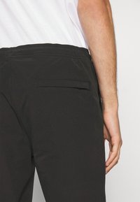 Černé kalhoty s hladkým povrchem, vybavené zadní kapsou a elastickým pasem. Pozadí je neutrální, zaměřené na detaily oděvu.