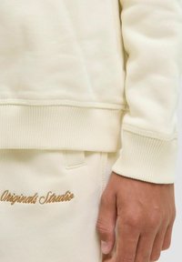 Sweatshirt de cor creme com punhos e barra canelados, tecido texturizado e bordado castanho em contraste que diz "Originals Studio."