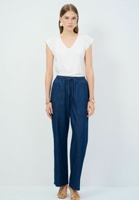 Camiseta blanca de cuello en V y pantalones vaqueros oscuros holgados con cintura con cordón. El modelo lleva sandalias doradas y pendientes decorativos. Diseño simple y casual.