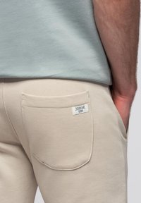 Beige sweatpants met een achterzak met een label, gecombineerd met een lichtblauw shirt, dat een zachte textuur en een ontspannen pasvorm toont.