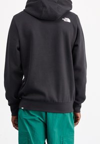 Uomo che indossa un hoodie nero The North Face e pantaloni cargo verdi, in piedi con le spalle rivolte verso la macchina fotografica su uno sfondo bianco.