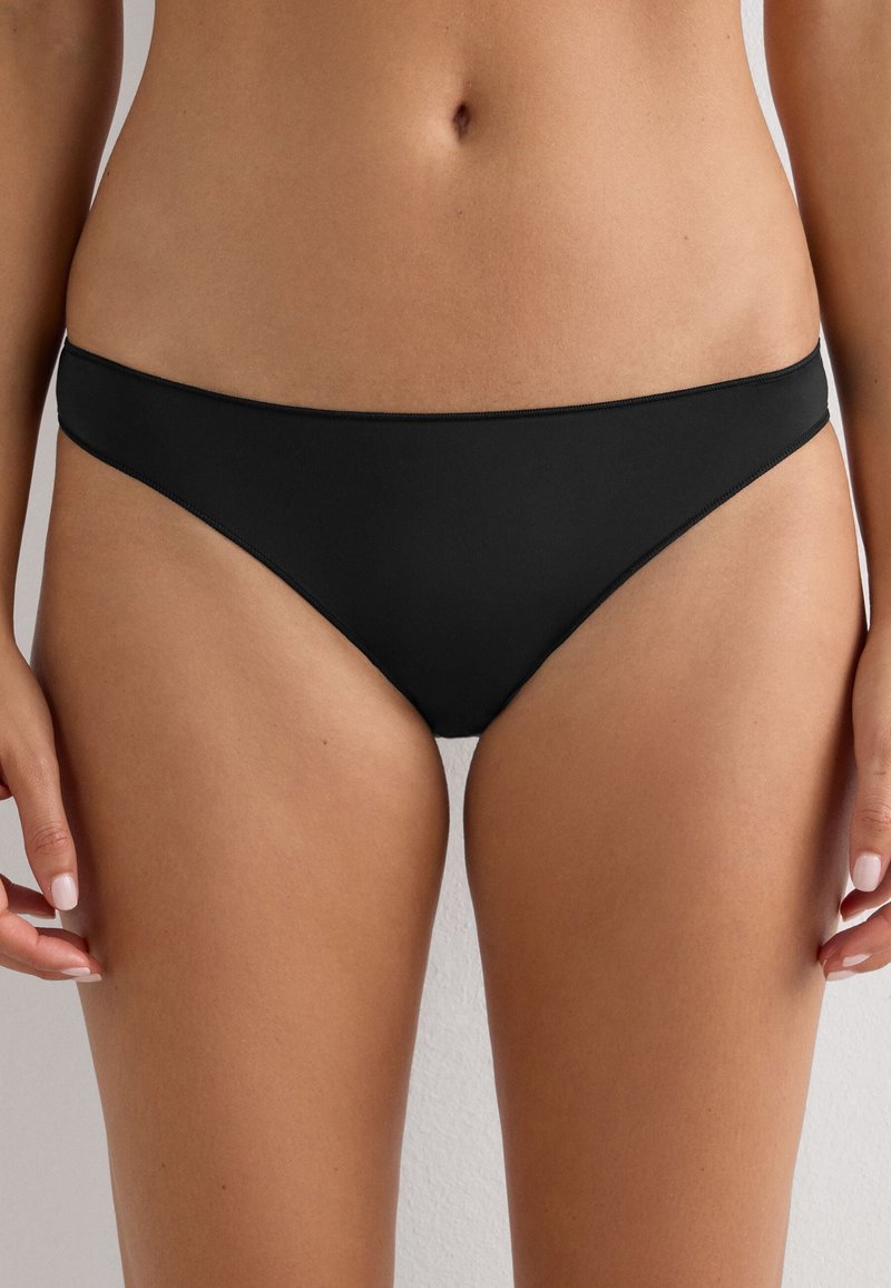 Kobieta nosząca proste czarne majtki w stylu bikini, pokazana od talii do górnej części ud na neutralnym tle.