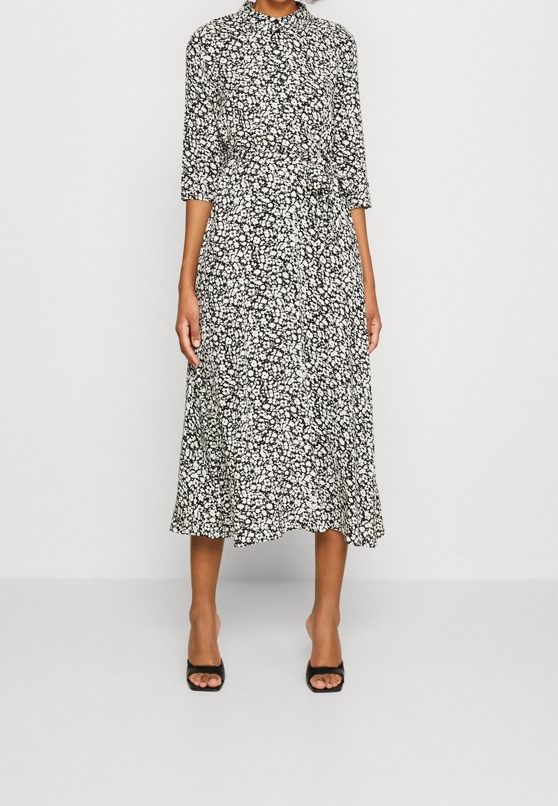 Femme portant une robe midi fleurie noire et blanche à manches trois-quarts et des escarpins noirs à bouts ouverts, debout devant un fond uni.