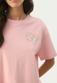 T-shirt en coton rose à manches courtes, avec un design floral brodé en marron sur le côté gauche de la poitrine.
