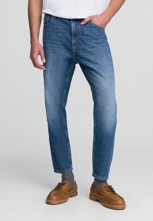 Jeans in denim blu con una vestibilità dritta, caratterizzati da cuciture arancioni, cinque tasche e una texture leggermente sbiadita. Indossati con scarpe marroni.