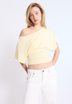 Blondínka v bledomodrom topu s odhalenými ramenami a bielych nohaviciach, stojaca so založenými rukami za chrbtom na jednofarebnom pozadí.