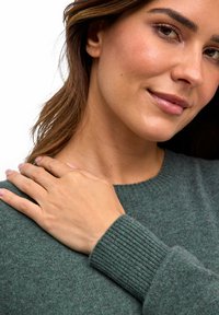 Maglione verde con polsini a coste, realizzato in materiale morbido. Inquadratura ravvicinata di una persona che poggia la mano sulla spalla.
