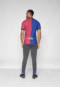 Heren voetbaltshirt, gesplitst rood en blauw ontwerp, korte mouwen, met UNHCR-logo. Draagt het met grijze leggings en zwarte voetbalschoenen.