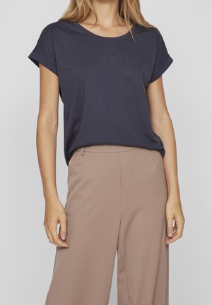 Vrouw met een marineblauwe blouse met korte mouwen, ingestopt in beige, hoog getailleerde, wijde broek tegen een effen achtergrond.