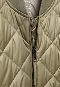 Veste matelassée vert olive avec fermeture éclair noire partiellement ouverte, montrant le col côtelé et l’étiquette intérieure.