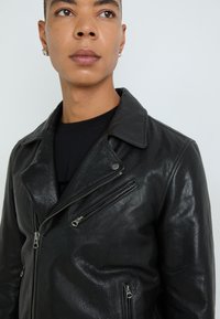 Must värvi nahast mootorrattajope tekstureeritud viimistlusega, laia krae, asümmeetriline tõmblukk ja mitmed tõmblukuga taskud.