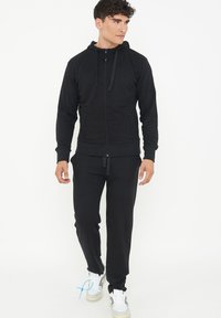 Felpa nera con zip e cappuccio, dotata di lacci, abbinata a pantaloni jogger neri. Realizzata in materiale morbido, presenta tasche laterali e polsini a coste.