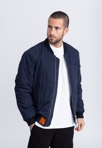 Marineblauwe bomberjack met een ritssluiting aan de voorkant, geribbelde manchetten en een oranje label op de zoom. Gemaakt van een gladde, gestructureerde stof.