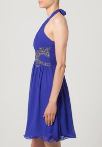 Halter-nack blå klänning med plisserad bodice, dekorerad med metalliska detaljer vid midjan, flytande chiffongkjol och en figurnära silhuett.