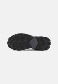 Semelle en caoutchouc noir avec un motif de semelle profonde, présentant des crampons inclinés pour une meilleure traction. Semelle intermédiaire grise partiellement visible avec une finition lisse.