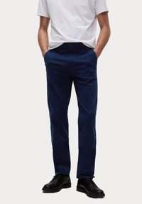 Marineblauwe chino's van katoen, met zijzakken, een knoopsluiting en een rechtlopende pasvorm. Gdragen met een wit T-shirt en zwarte schoenen.