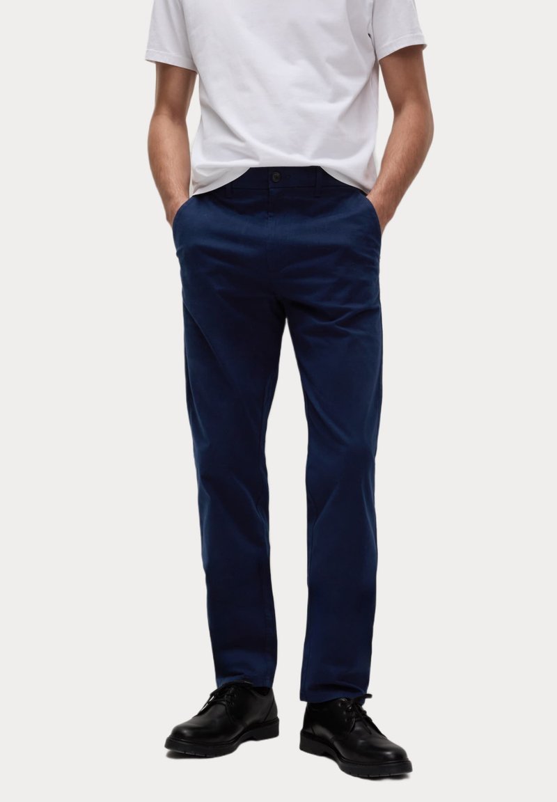 Marineblauwe chino's van katoen, met zijzakken, een knoopsluiting en een rechtlopende pasvorm. Gdragen met een wit T-shirt en zwarte schoenen.