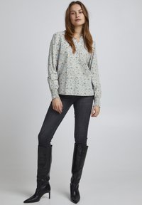 Blumige Bluse mit langen Ärmeln in Hellblau, mit Kragen und elastischen Bündchen, kombiniert mit dunklen Skinny-Jeans und schwarzen knielangen Stiefeln.