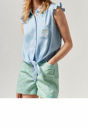 Mujer que lleva una camisa sin mangas azul claro con botones y un nudo en el delantero y un bolsillo bordado con flores, combinada con pantalones cortos florales en verde menta.