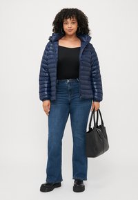 Dunkelblaue Steppjacke mit Reißverschluss vorne, getragen über einem schwarzen Oberteil. Kombiniert mit hochgeschnittenen Jeans und einer schwarzen Handtasche. Mattes Material, lässige Passform.