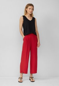 Top nero senza maniche con scollo a V, abbinato a pantaloni rossi larghi con cintura elastica e tasche, indossato con sandali neri.
