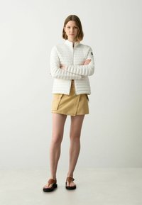 Witte, gequiltte zip-up jas met een hoge kraag, gecombineerd met een beige rok met cargozakken en een paar bruine sandalen.