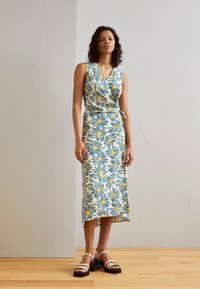 Robe portefeuille florale avec un décolleté en V, présentant un motif de fleurs bleues et jaunes sur un fond clair. Associée à des sandales à talons avec lanières.