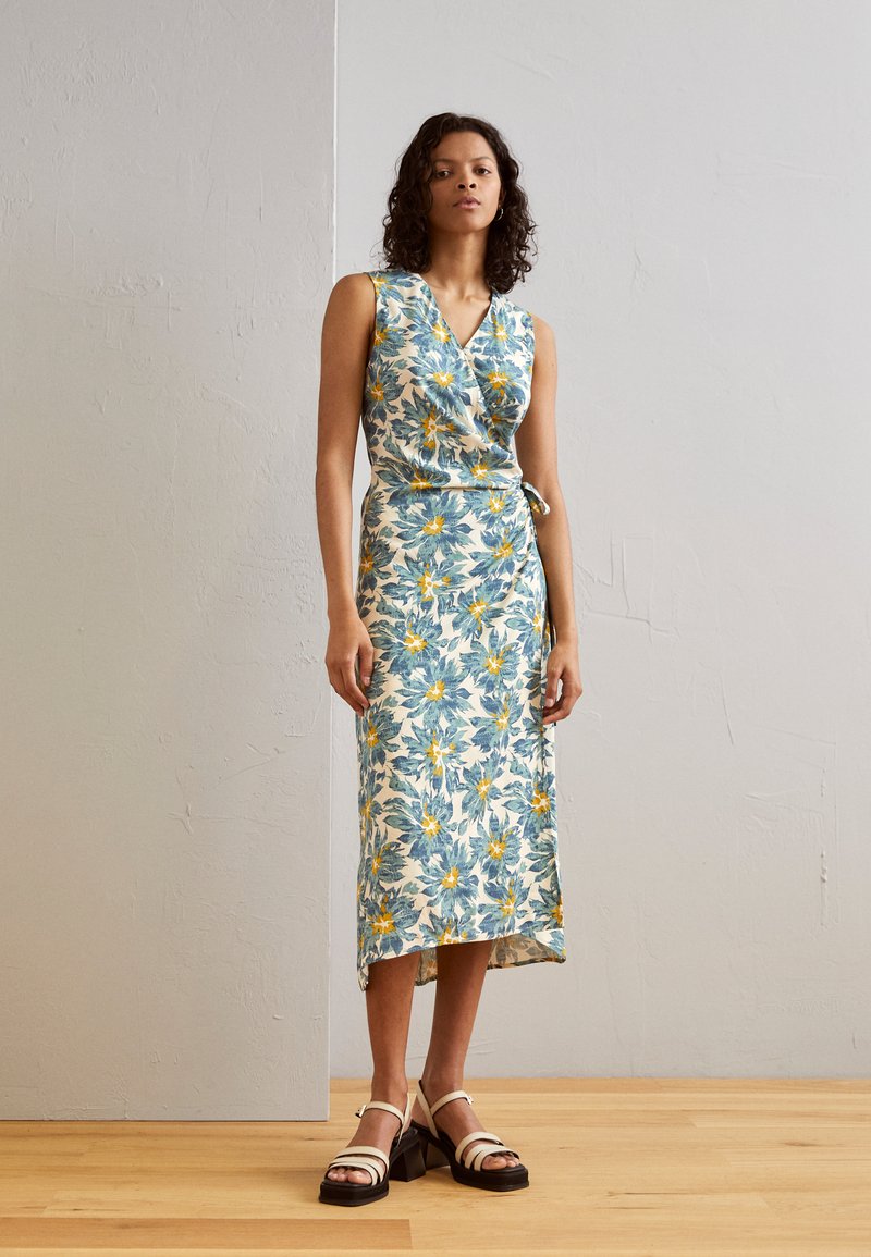 Robe portefeuille florale avec un décolleté en V, présentant un motif de fleurs bleues et jaunes sur un fond clair. Associée à des sandales à talons avec lanières.