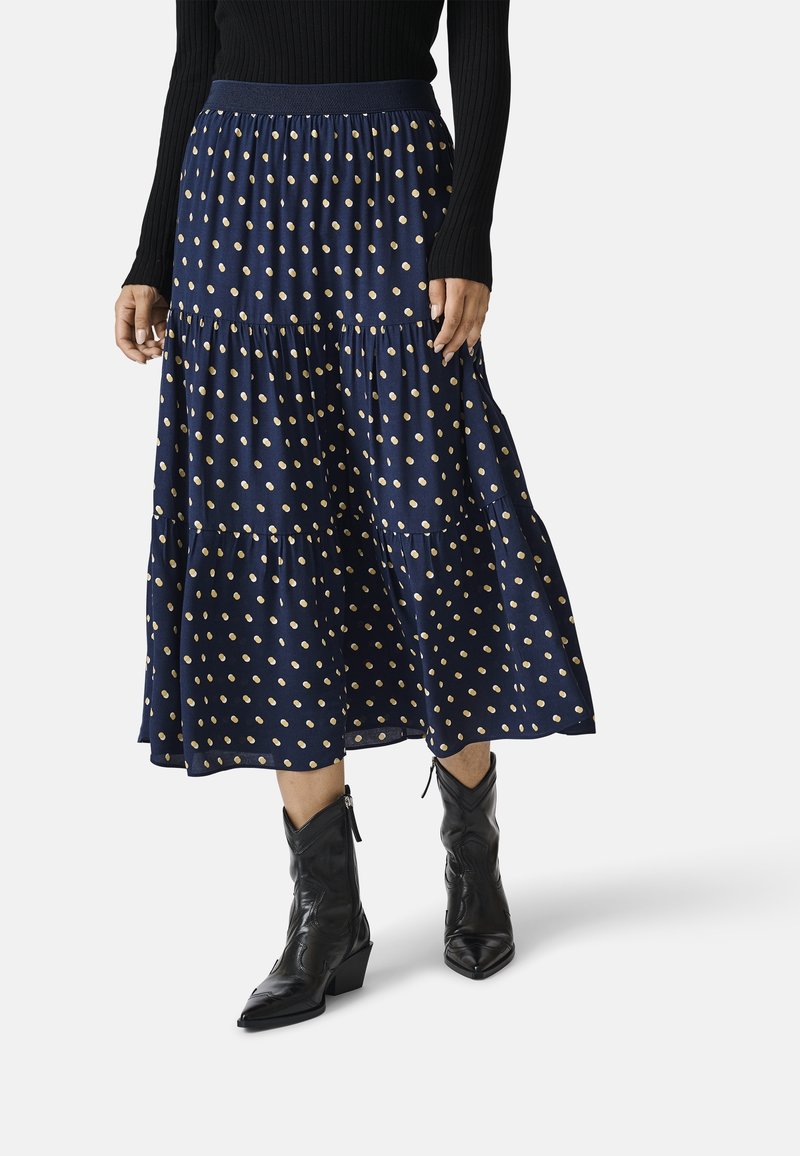 Personne portant une jupe midi bleu marine à pois beige, un haut noir à manches longues et des bottines noires à bout pointu.