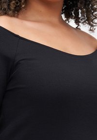 Nainen, jolla on päällään musta ribbattu kyynärpään yli ulottuva paita, paljastaen dekolteen ja kiharat hiukset yksinkertaista taustaa vasten.