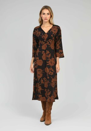 Une femme se tient debout, portant une robe midi noire avec un imprimé floral marron et des manches trois-quarts, associée à des bottes marron en daim montantes.
