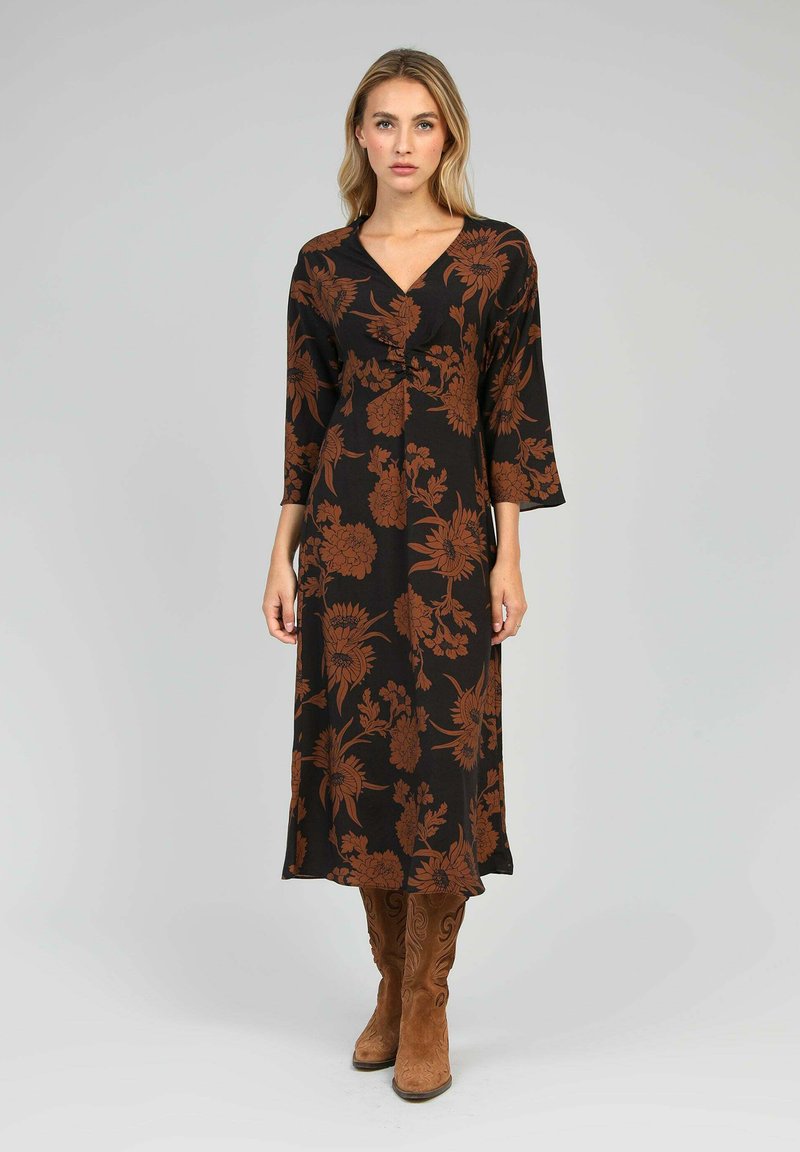 Une femme se tient debout, portant une robe midi noire avec un imprimé floral marron et des manches trois-quarts, associée à des bottes marron en daim montantes.