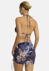 Se muestra a una mujer de espaldas, llevando un bikini de estampado floral y una cubierto transparente a juego, estilizado con un cuello halter atado.
