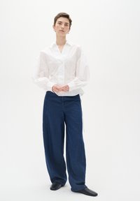 Camicia bianca a maniche lunghe con colletto e maniche a sbuffo, abbinata a jeans larghi blu scuro. Scarpe nere completano l'outfit.