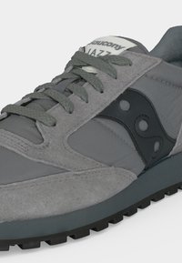 Γκρι αθλητικό παπούτσι Saucony Jazz με σκούρα γκρι κορδόνια, μαύρο λογότυπο στο πλάι, υφασμένο πέλμα και λευκή ετικέτα στη γλώσσα.