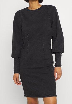 Kvinde iført en mørkegrå, tætsiddende knælængde kjole med puffede lange ærmer og rynkede manchetter, stående mod en ensfarvet baggrund.