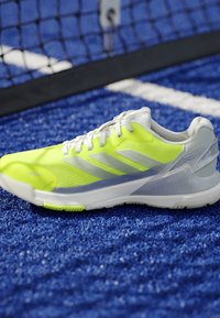 Bright yellow and white athletic shoe on a blue textured sports court near a black net with white lines.