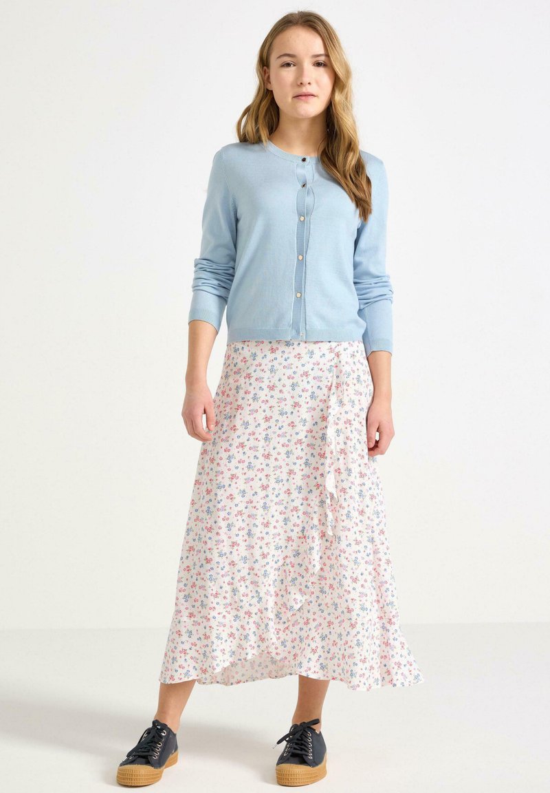 Lyseblå strikket cardigan med knapper, båret over en svajende hvid nederdel med blomsterprint i pink og blå, kombineret med sorte sko.