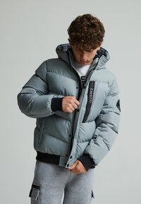 Doudoune grise avec capuche, présentant une fermeture éclair avant et une poche estampillée, des poignets élastiques, et un design matelassé.