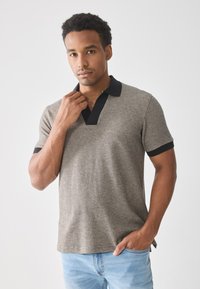 Kortermet polo skjorte i beige med et teksturert mønster. Svart krage og mansjetter gir kontrast. Modellen holder kragen med den ene hånden.