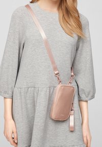 Różowo-złota torebka crossbody wykonana z gładkiego materiału, o prostokątnym kształcie, z regulowanym paskiem i małym uchwytem na nadgarstek.