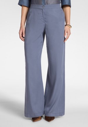 Femme portant un pantalon bleu taille haute à jambes larges, des chaussures marron pointues et un chemisier foncé avec un bracelet au poignet droit.